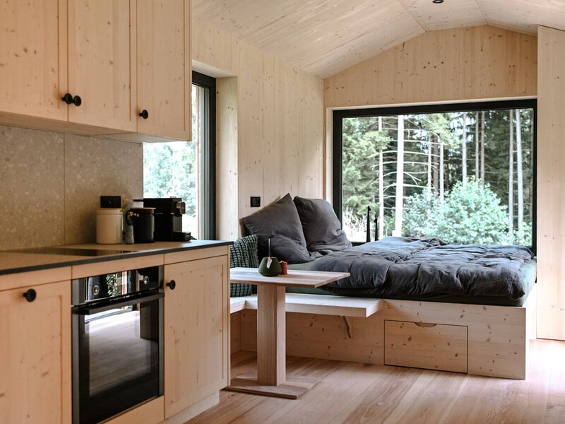 Ein gemütlicher Wohnbereich mit einer modernen Holz-Kitchenette und einem einladenden Bett direkt am großen Panoramafenster mit Waldblick.