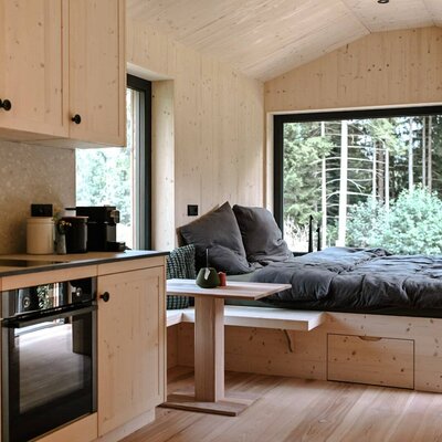 Ein gemütlicher Wohnbereich mit einer modernen Holz-Kitchenette und einem einladenden Bett direkt am großen Panoramafenster mit Waldblick.