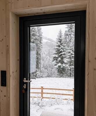 Blick durch die moderne Glastür auf eine idyllische, verschneite Winterlandschaft mit dichten Nadelbäumen.