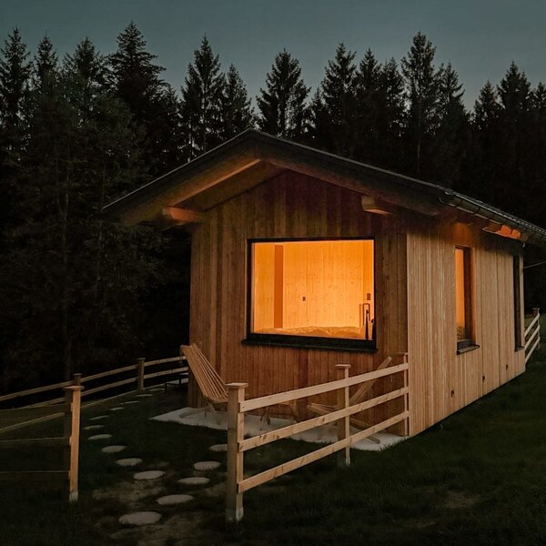 Ein gemütliches, warm beleuchtetes Holzhaus mit einer kleinen, umzäunten Terrasse direkt am ruhigen Waldrand.