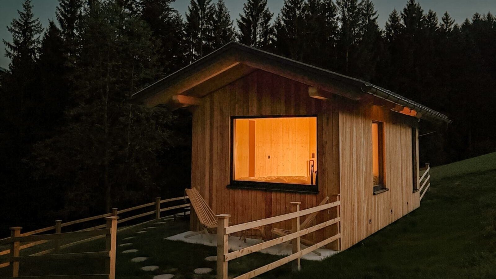 Ein gemütliches, warm beleuchtetes Holzhaus mit einer kleinen, umzäunten Terrasse direkt am ruhigen Waldrand.