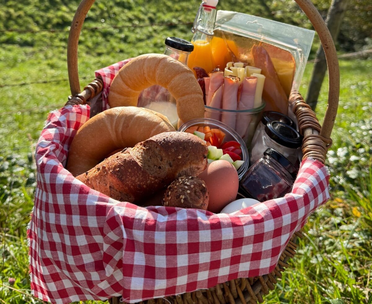 Ein reichhaltiger Frühstückskorb des Bauernhofs mit frischen Backwaren, herzhaften Spezialitäten und Säften auf einer grünen Wiese.