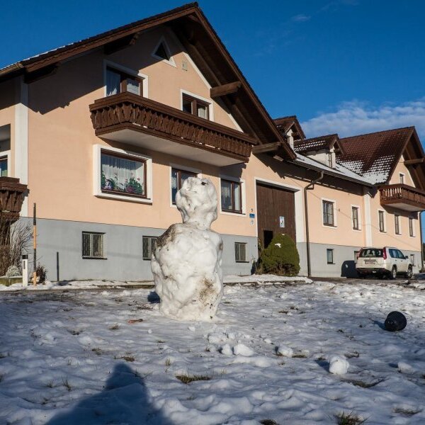 Die Außenansicht des Bauernhofs im Winter mit verschneitem Vorgarten und Holzbalkonen.