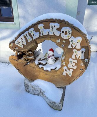 Willkommensschild aus Holz mit Schnee und einem Schneemann vor dem Bauernhof.