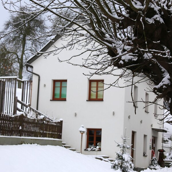 Der weiße Bauernhof in winterlicher Umgebung mit Schnee, Bäumen und einem Terrassenbereich mit Holzzaun.