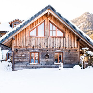Der Bauernhof im Winter, mit einer Holzfassade, Schnee und einer Bergkulisse.