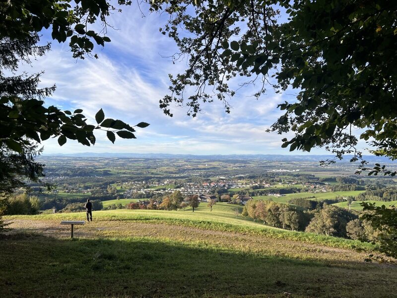 Panoramablick über die grüne Landschaft und eine Ortschaft in der Ferne, mit einer Bank und einer Person, die die Aussicht genießt.