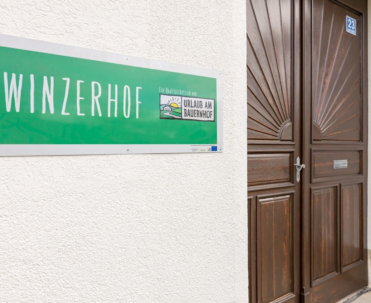 Das grüne Schild 'Winzerhof' und 'Urlaub am Bauernhof' neben der Eingangstür des Hauses mit der Hausnummer 23.