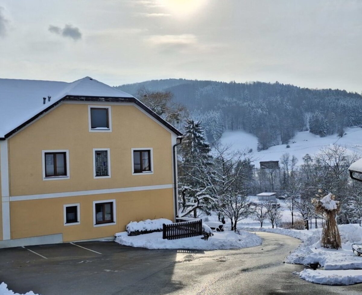 Der Bauernhof mit seinem gelben Hauptgebäude und einem Nebengebäude, umgeben von einer schneebedeckten Winterlandschaft.