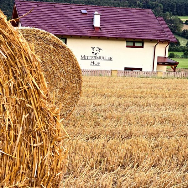 Der Bauernhof Mittermüller Hof mit rotem Dach, umgeben von Feldern und Heuballen im Vordergrund.