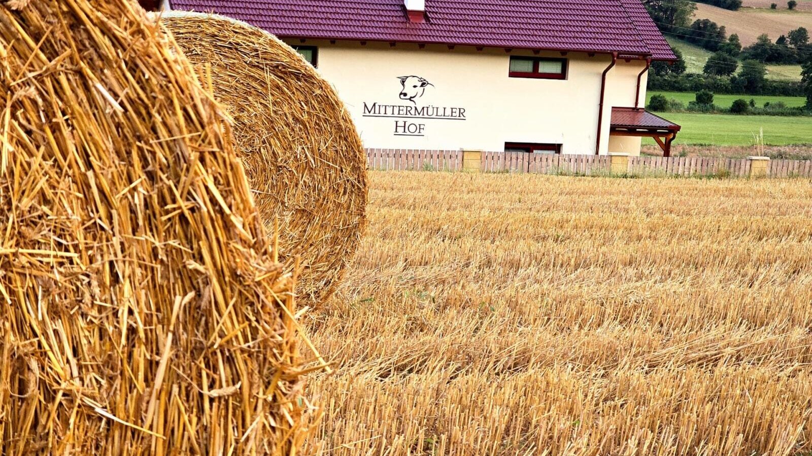 Der Bauernhof Mittermüller Hof mit rotem Dach, umgeben von Feldern und Heuballen im Vordergrund.