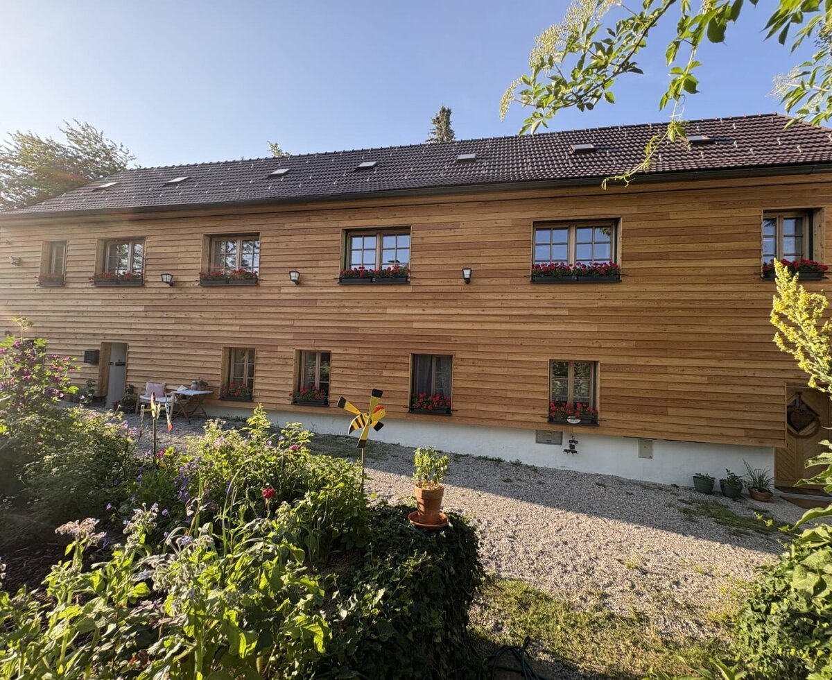 Außenansicht des Landhofs mit Holzfassade, Fenstern mit Blumenkästen und einem Gartenbereich.