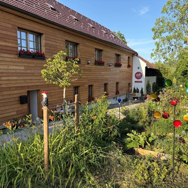 Außenansicht des Landhofs mit Holzfassade, Blumenkästen und einem Garten mit Pflanzen und einem bunten Windspiel.