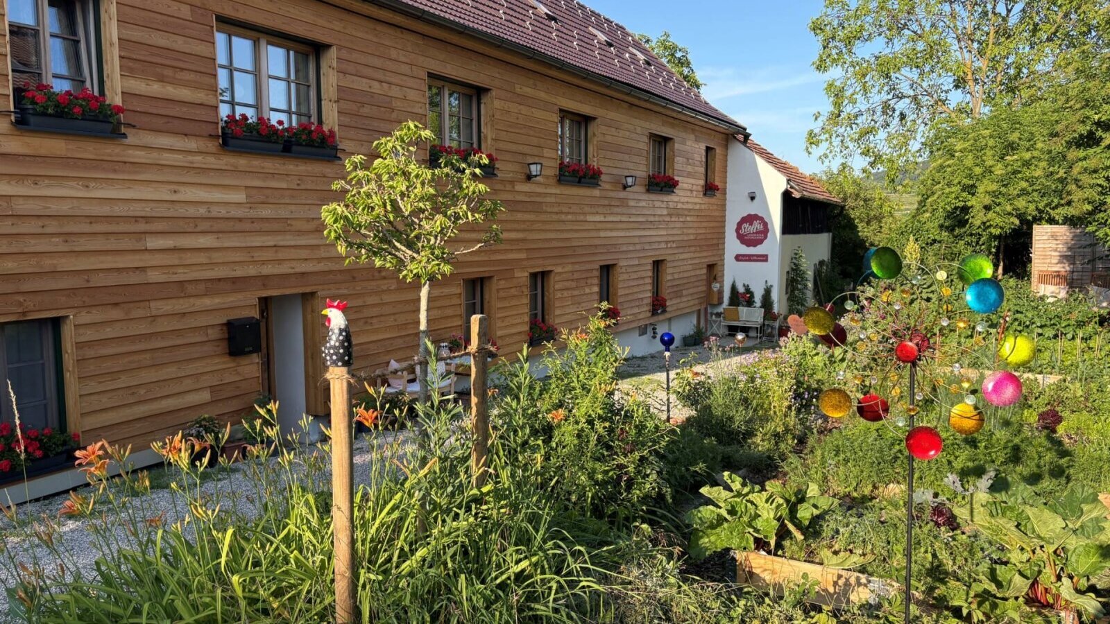 Außenansicht des Landhofs mit Holzfassade, Blumenkästen und einem Garten mit Pflanzen und einem bunten Windspiel.