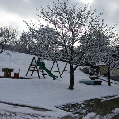 Der verschneite Außenbereich des Bauernhofs mit Kinderspielplatz, Rutsche, Schaukeln und einer Sitzgruppe aus Holz.