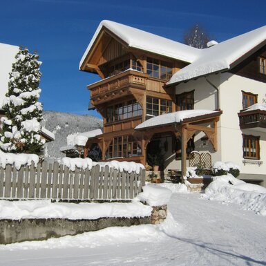 Außenansicht des schneebedeckten Bauernhofs mit Holzbalkonen und der Zufahrt im Winter.