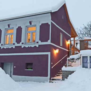 Die Fassade des Hofs im Winter, mit Schnee bedeckt, beleuchteten Fenstern und Außenzugang zu den Zimmern.