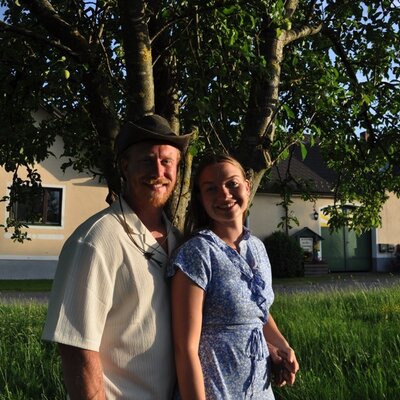 Zwei lächelnde Personen stehen vor dem Bauernhof, mit einem Baum und Gras im Vordergrund.