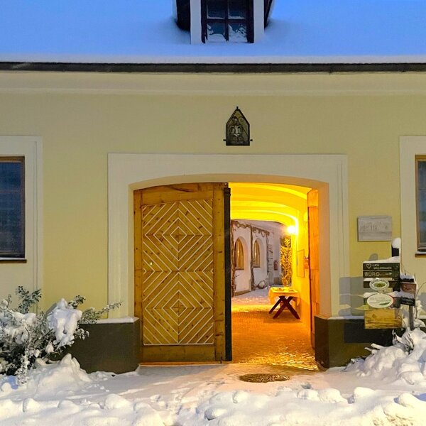 Der Eingang zum Hof mit offener Holztür und einem hell beleuchteten Durchgang, umgeben von Schnee.