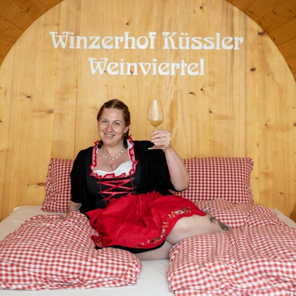 Eine der Weinfass-Suiten auf dem Winzerhof Küssler mit einem Doppelbett und einer Person, die ein Weinglas hält.