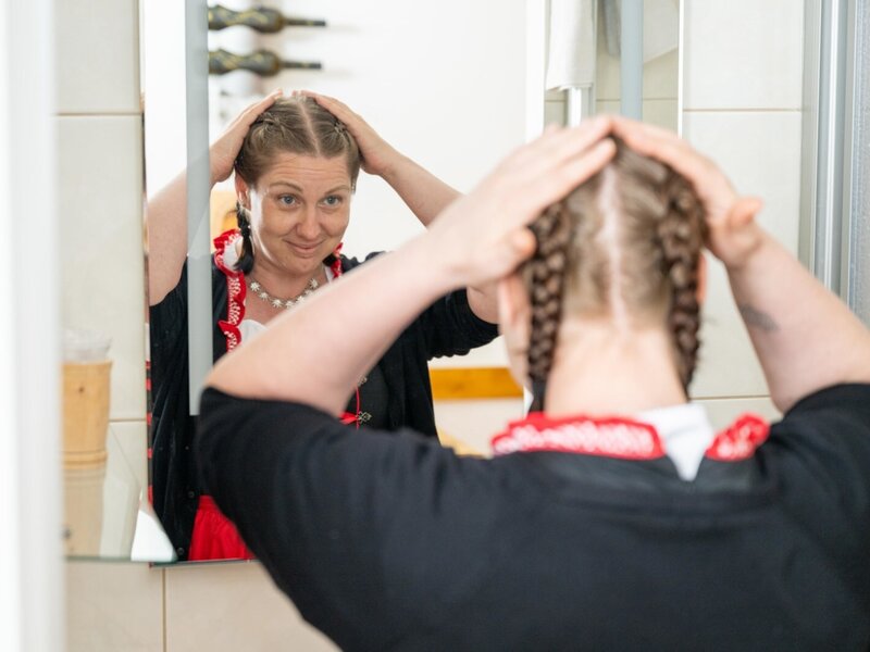 Eine Frau in traditioneller Kleidung blickt im Hof in den Spiegel und richtet ihre geflochtenen Haare, mit Weinflaschen im Hintergrund.