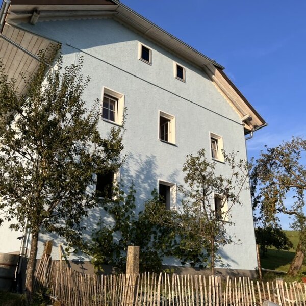 Außenansicht des Hofs mit heller Fassade, umgeben von Bäumen und einem Holzzaun.