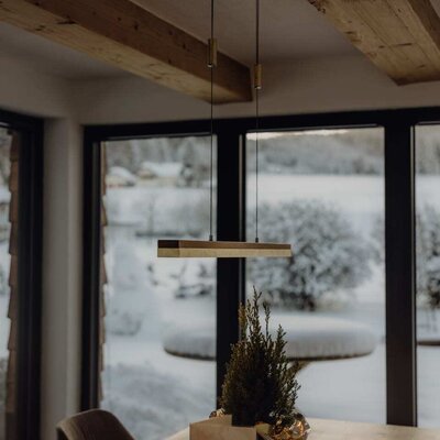 Essbereich im Bauernhof mit Holztisch, moderner Hängelampe, Holzbalken und großen Fenstern mit Blick auf die verschneite Landschaft.