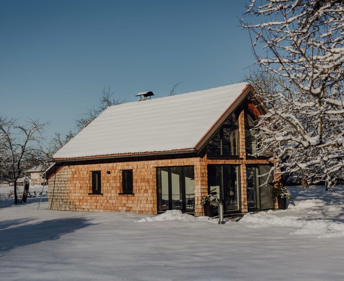 Das Chalet im Obstgarten des Bauernhofs mit schneebedecktem Dach und Holzfassade, umgeben von verschneiten Obstbäumen.