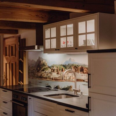 Die Küche des Bauernhofs ist ausgestattet mit weißen Schränken, dunkler Arbeitsplatte, Spüle, integriertem Backofen und Kochfeld, beleuchteten Oberschränken und einer Wandverkleidung mit Landschaftsmotiv.