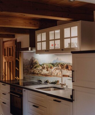 Die Küche des Bauernhofs ist ausgestattet mit weißen Schränken, dunkler Arbeitsplatte, Spüle, integriertem Backofen und Kochfeld, beleuchteten Oberschränken und einer Wandverkleidung mit Landschaftsmotiv.