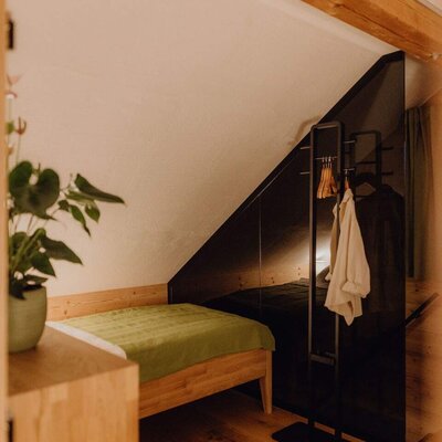 Schlafzimmer im Bauernhof mit Einzelbett, Holzboden, Dachschräge, Holzbalken und einem dunklen Raumteiler mit Kleiderständer.