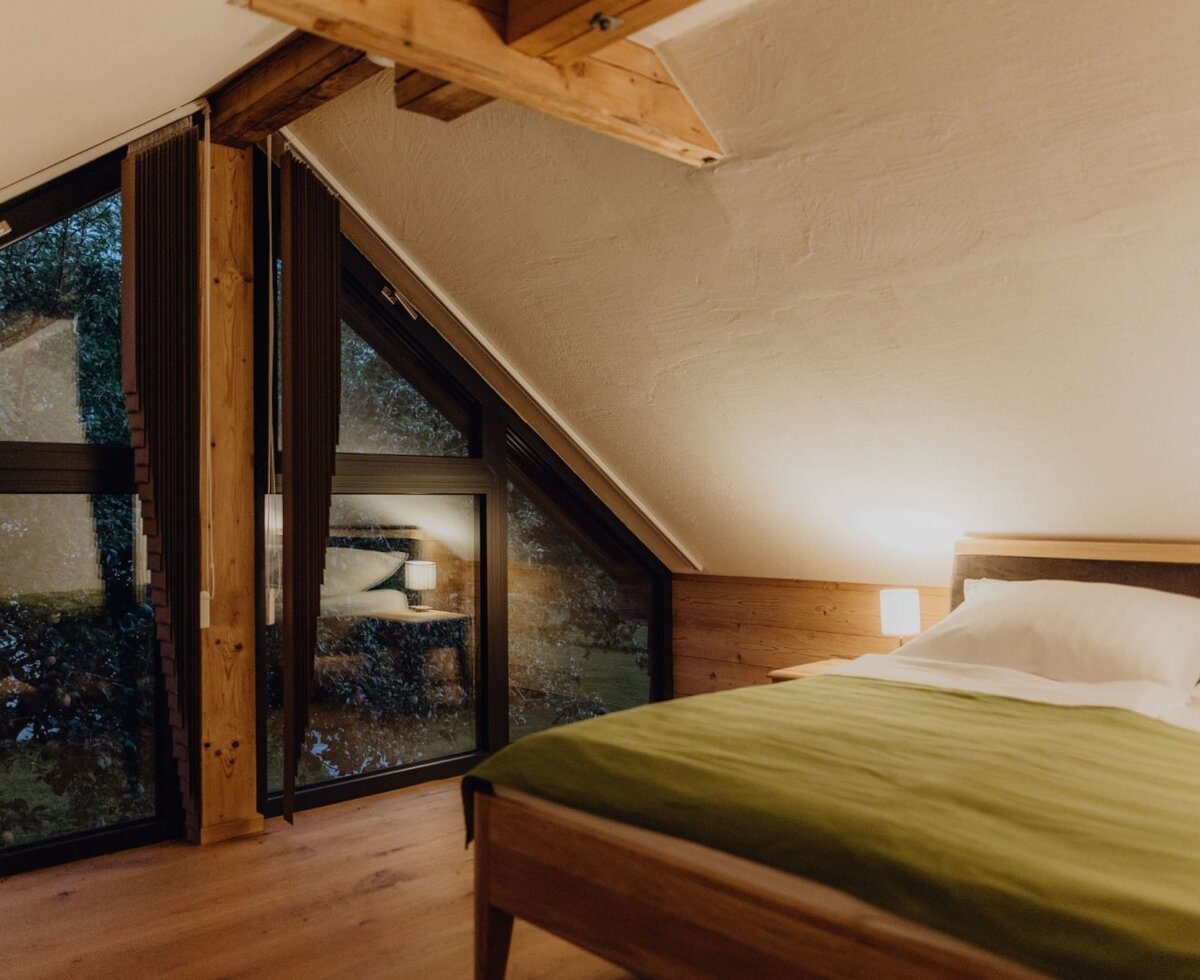 Schlafzimmer im Bauernhof mit Doppelbett, Holzelementen und großen Fenstern mit Blick ins Grüne.