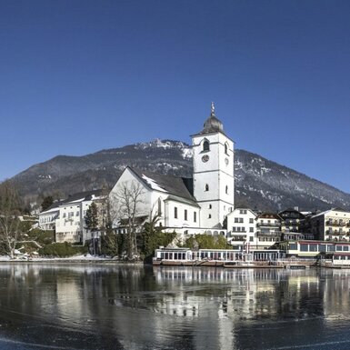 Der Wolfgangsee im Winter mit der Wallfahrtskirche St. Wolfgang und den schneebedeckten Bergen des Salzkammergutes.