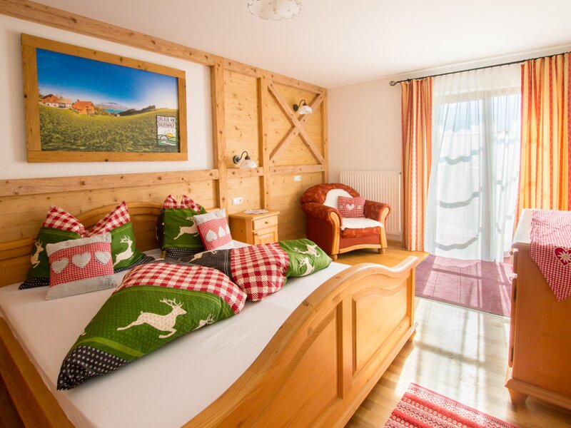 Doppelzimmer im Bauernhof mit Holzbett, traditioneller Bettwäsche, Sessel und direktem Zugang zum Balkon.