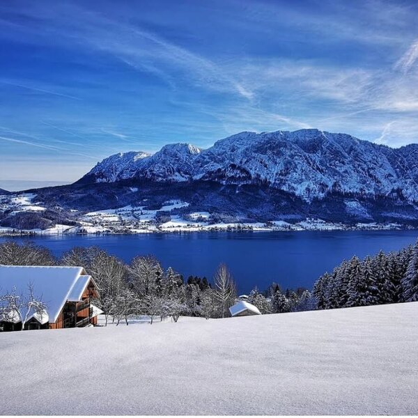 Winterlandschaft mit einem Bauernhof, dem Attersee und dem schneebedeckten Höllengebirge.