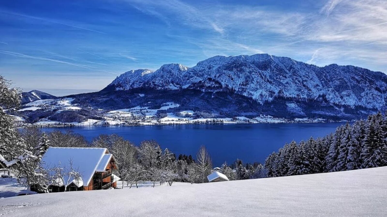 Winterlandschaft mit einem Bauernhof, dem Attersee und dem schneebedeckten Höllengebirge.