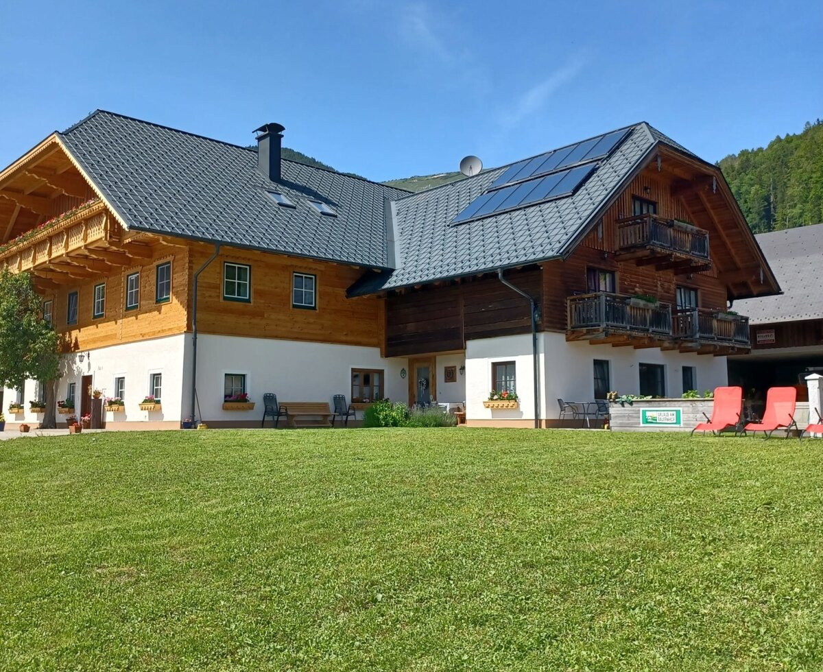 Der Bauernhof mit traditioneller Holz- und Putzfassade, Balkonen, Solaranlage und einer Liegewiese mit roten Sonnenliegen.