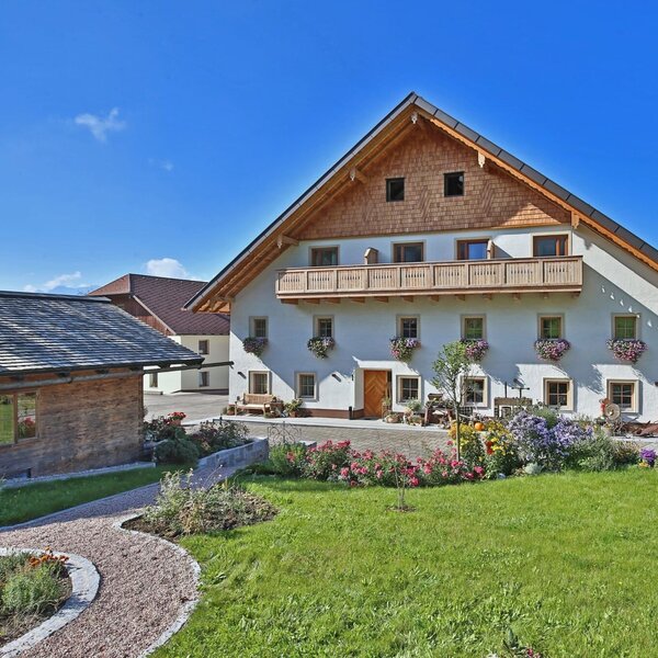 Das Hauptgebäude des Bauernhofs mit Holzbalkon, Blumenkästen und einem Vorgarten mit Rasenfläche und Kiesweg.
