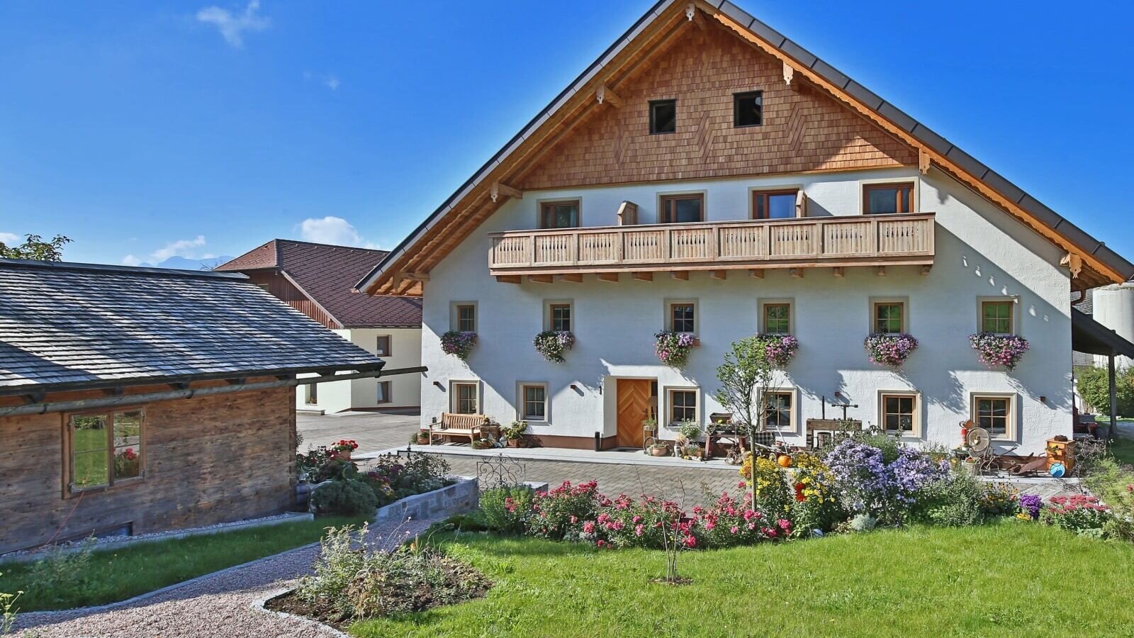 Das Hauptgebäude des Bauernhofs mit Holzbalkon, Blumenkästen und einem Vorgarten mit Rasenfläche und Kiesweg.