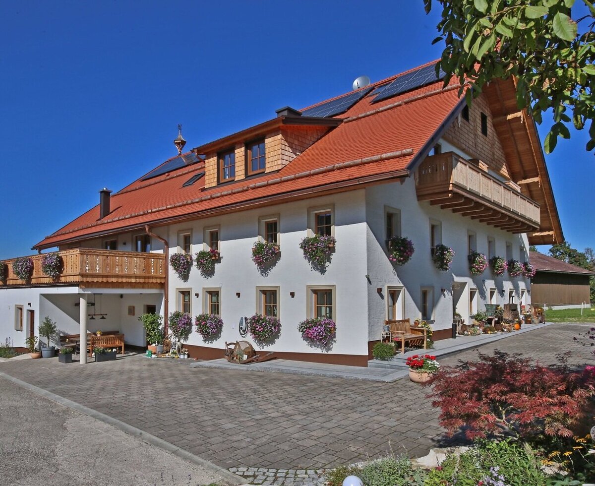 Außenansicht des Bauernhofs mit weißen Wänden, Holzbalkonen, Blumenkästen und gepflasterter Zufahrt.
