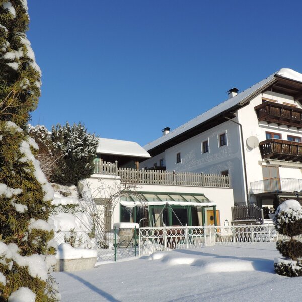 Das Bauernhof-Gebäude mit Holzbalkonen und einem schneebedeckten Garten unter klarem blauem Himmel.