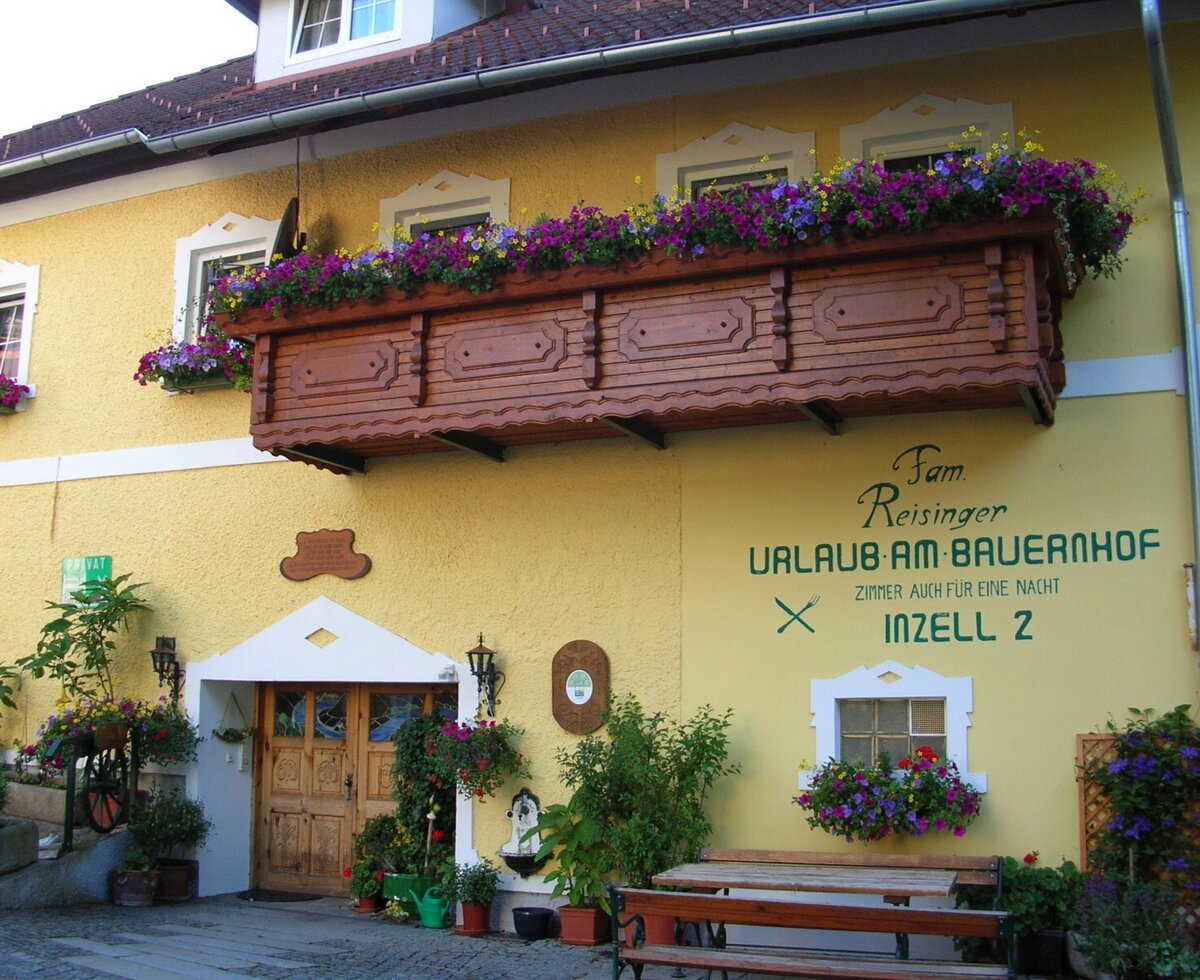 Die Fassade des Bauernhofs mit traditionellem Holzbalkon, Blumenkästen und einem einladenden Eingangsbereich mit Außensitzgelegenheit.