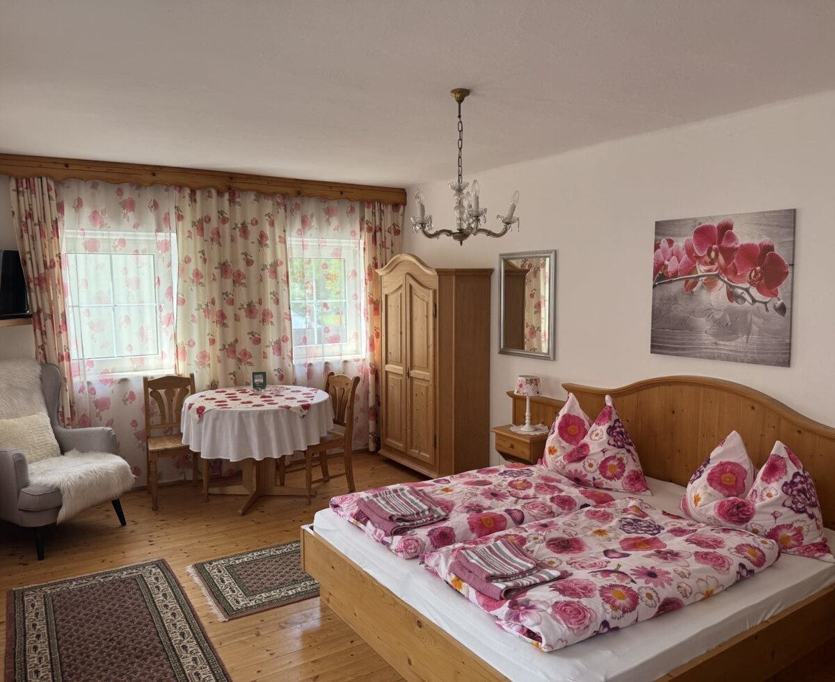 Doppelzimmer im Bauernhof mit Holzbett, Kleiderschrank, Tisch, zwei Stühlen und zwei Fenstern.