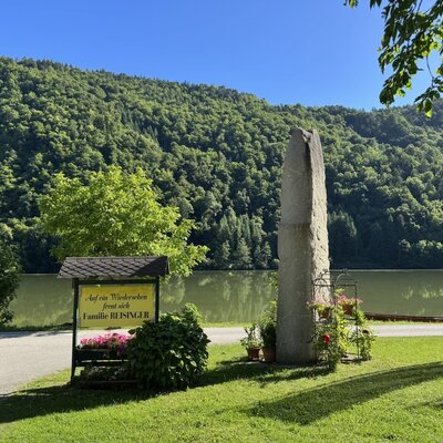 Der Außenbereich des Bauernhofs mit einem Schild der Familie Reisinger, einem Steinmonument und Blick auf den See und die bewaldeten Berge.