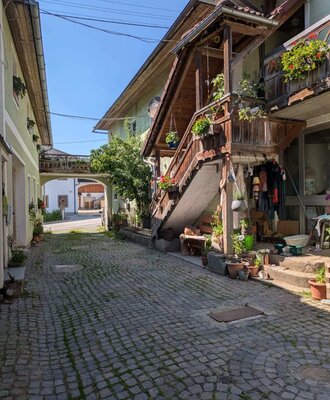 Der gepflasterte Weg zwischen den Gebäuden des Bauernhofs, mit einer Außentreppe und blumengeschmückten Balkonen.