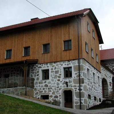 Die Außenansicht des Bauernhofs mit Steinsockel, Holzfassade und überdachtem Terrassenbereich.