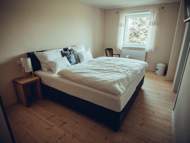 Schlafzimmer im Bauernhof mit Doppelbett, Nachttisch mit Lampe, Holzboden und einem Fenster mit Vorhängen.