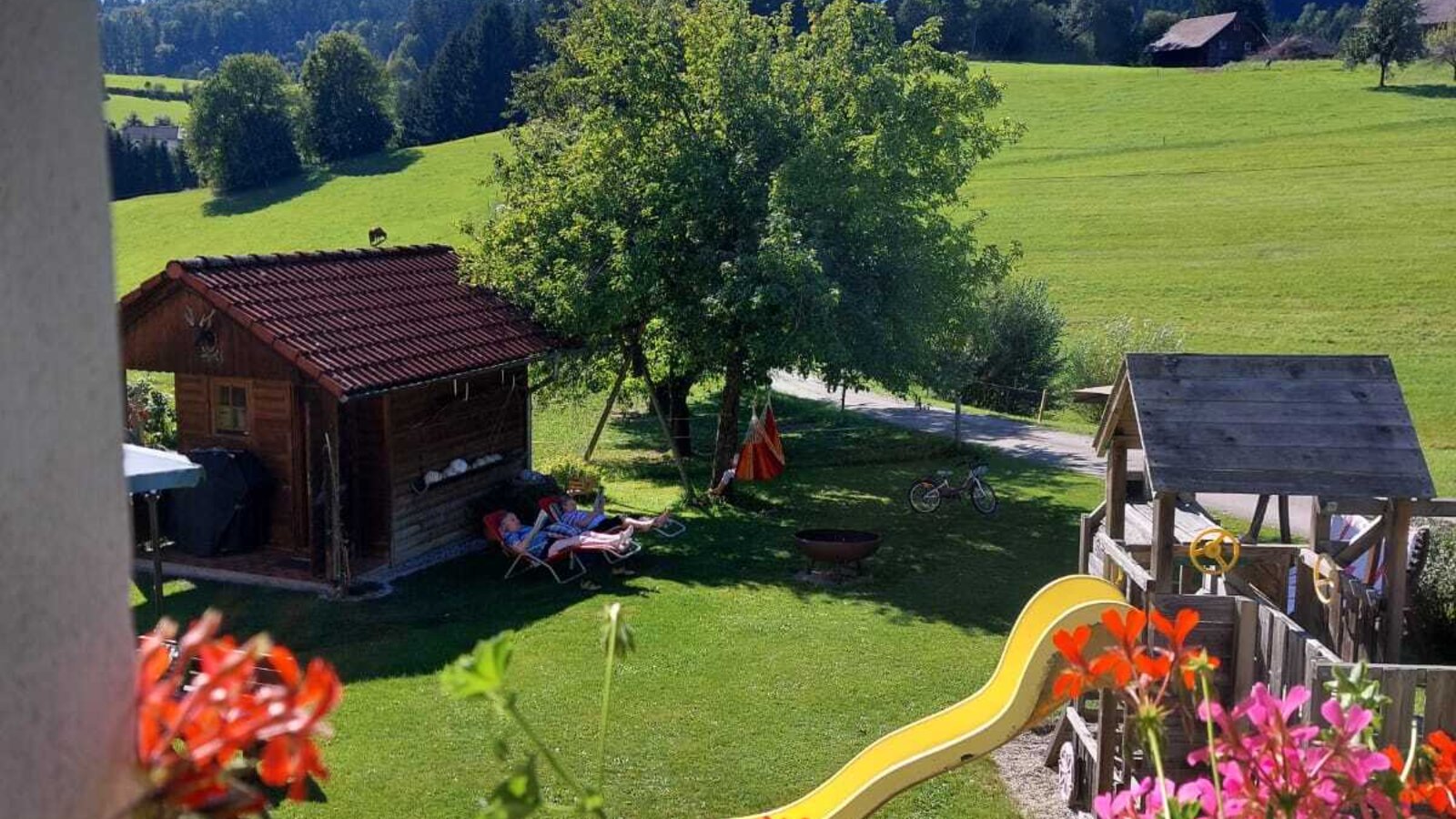 Blick vom Bauernhof auf den Gartenbereich mit Rutsche und Spielhaus vor der Kulisse von Wiesen und Wäldern.