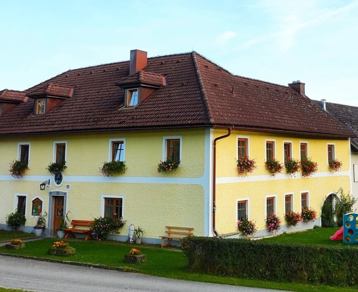 Der Bauernhof mit gelber Fassade, rotem Ziegeldach, Blumenkästen, einem Kinderspielplatz und Parkplätzen.