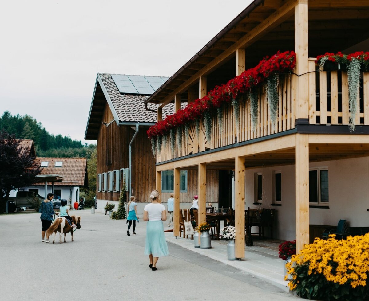 Die Hofanlage des Bauernhofs mit blumengeschmückten Holzbalkonen, Solaranlagen auf dem Dach und dem Hofplatz, wo Gäste und Ponys unterwegs sind.